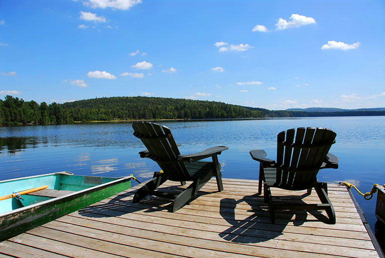 Two adirondack chairs set on the edge of a dock overlooking a lake, with green trees and blue skies in the background.
